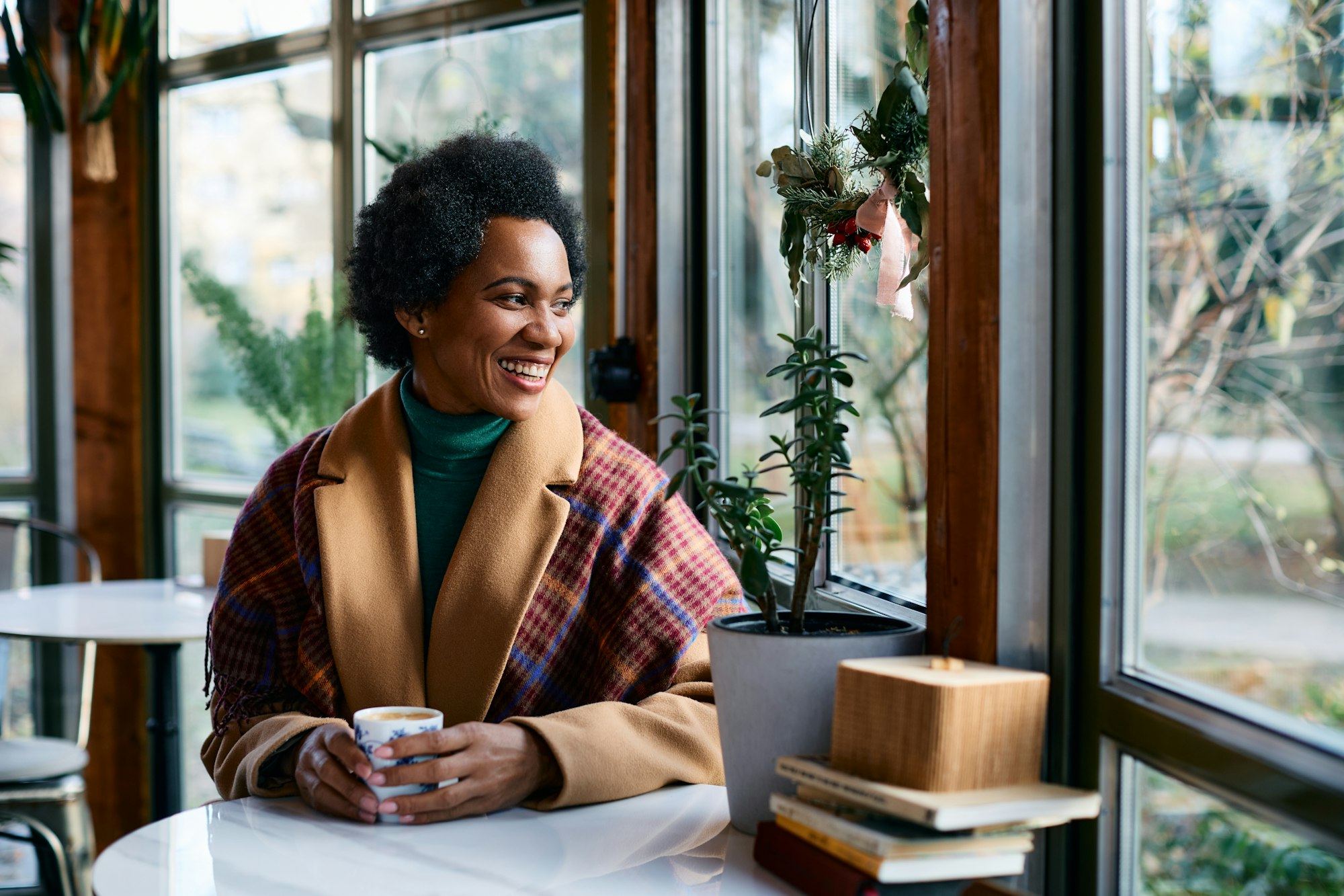 Happy black woman drinking coffee in cafe and looking through the widow.